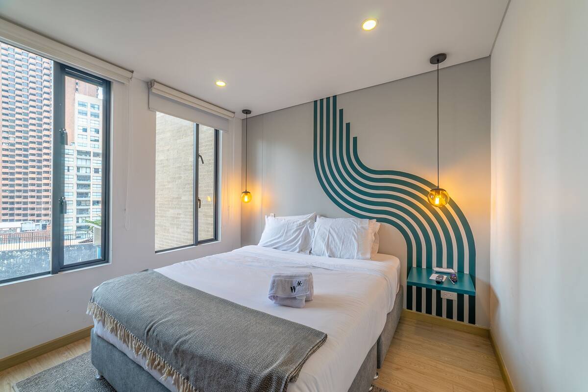 A modern hotel room with a large bed, a view of the city through a window, and a unique wall design.