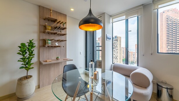 Una sala de estar moderna con una mesa de comedor de vidrio, sillas blancas y negras, una lámpara colgante y vistas de la ciudad a través de amplios ventanales.