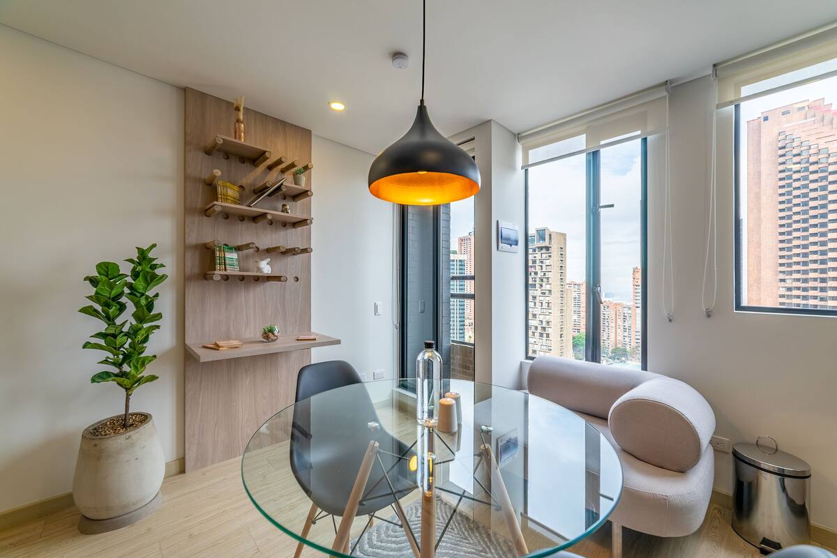 A modern living room with a glass dining table, black and white chairs, a hanging pendant light, and a view of the city through large windows.
