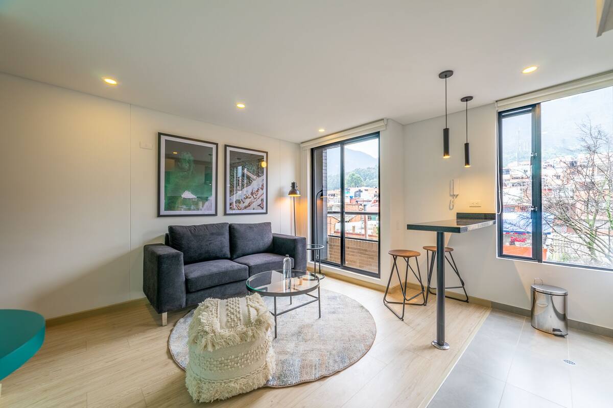 A modern living room with a grey sofa, a glass coffee table, and a round rug. There are two framed pictures on the wall, a large window with a city view, and a small table with stools.