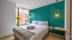 Una habitación de hotel moderna con una pared de acento color turquesa, una cama grande con ropa de cama blanca, una mesita de noche con lámpara y vistas a la ciudad a través de un ventanal amplio.