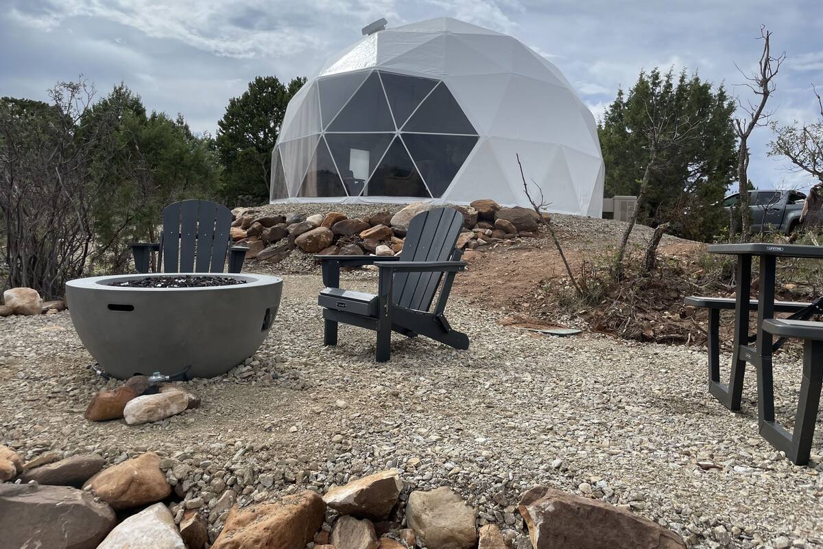 A geodesic dome structure with a fire pit and Adirondack chairs in front.