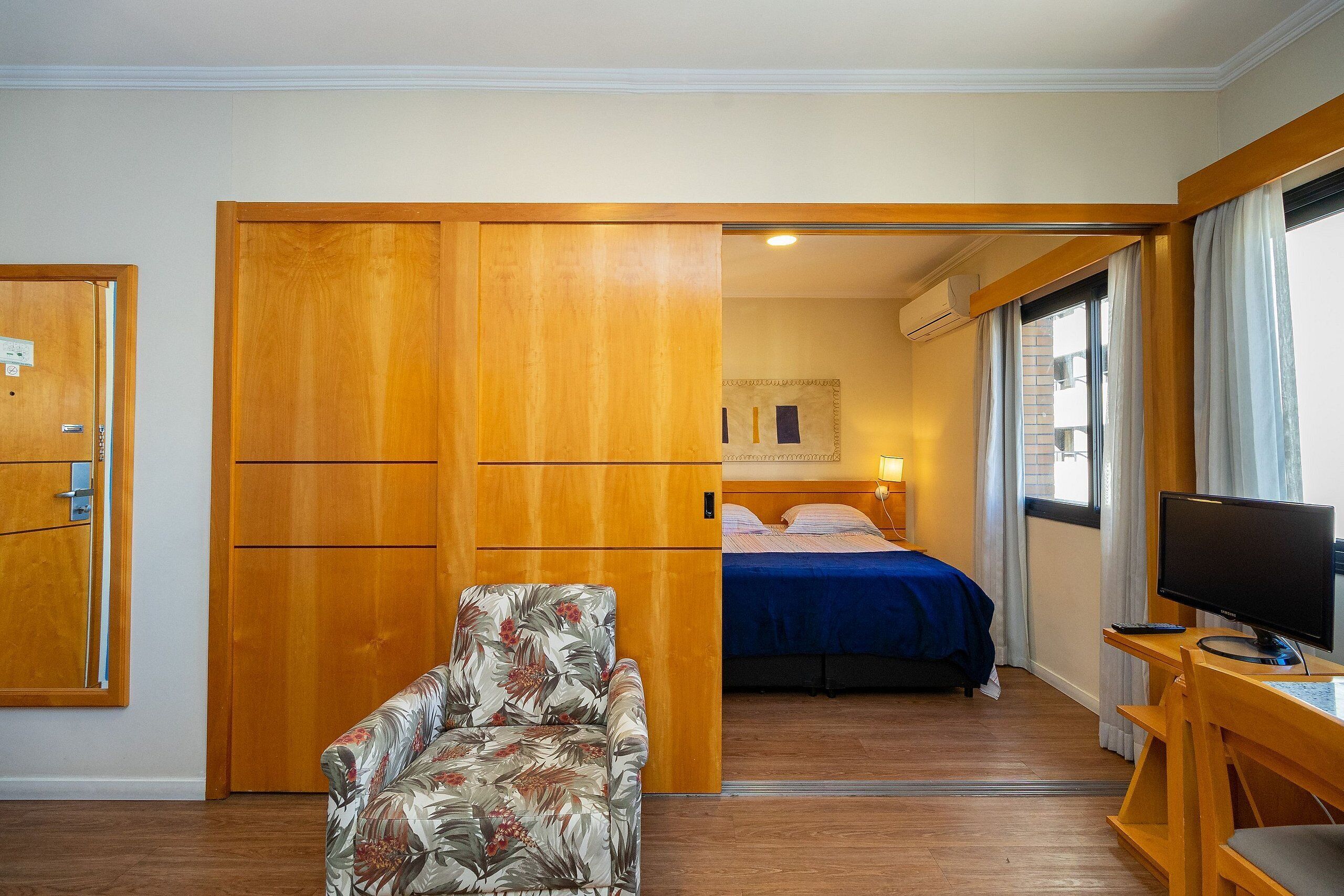 A hotel room with a bed, a desk with a computer, a chair, and a wardrobe.