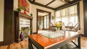 Une salle à manger avec une table en bois dressée pour un repas, entourée de chaises. Une cheminée avec une manteau de cheminée est visible sur la gauche, et une fenêtre avec des rideaux se trouve sur la droite.