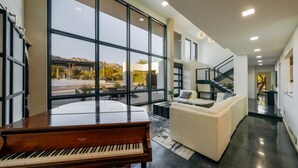 Un amplio salón con un piano de cola, un sofá blanco y grandes ventanales que ofrecen vistas al exterior.