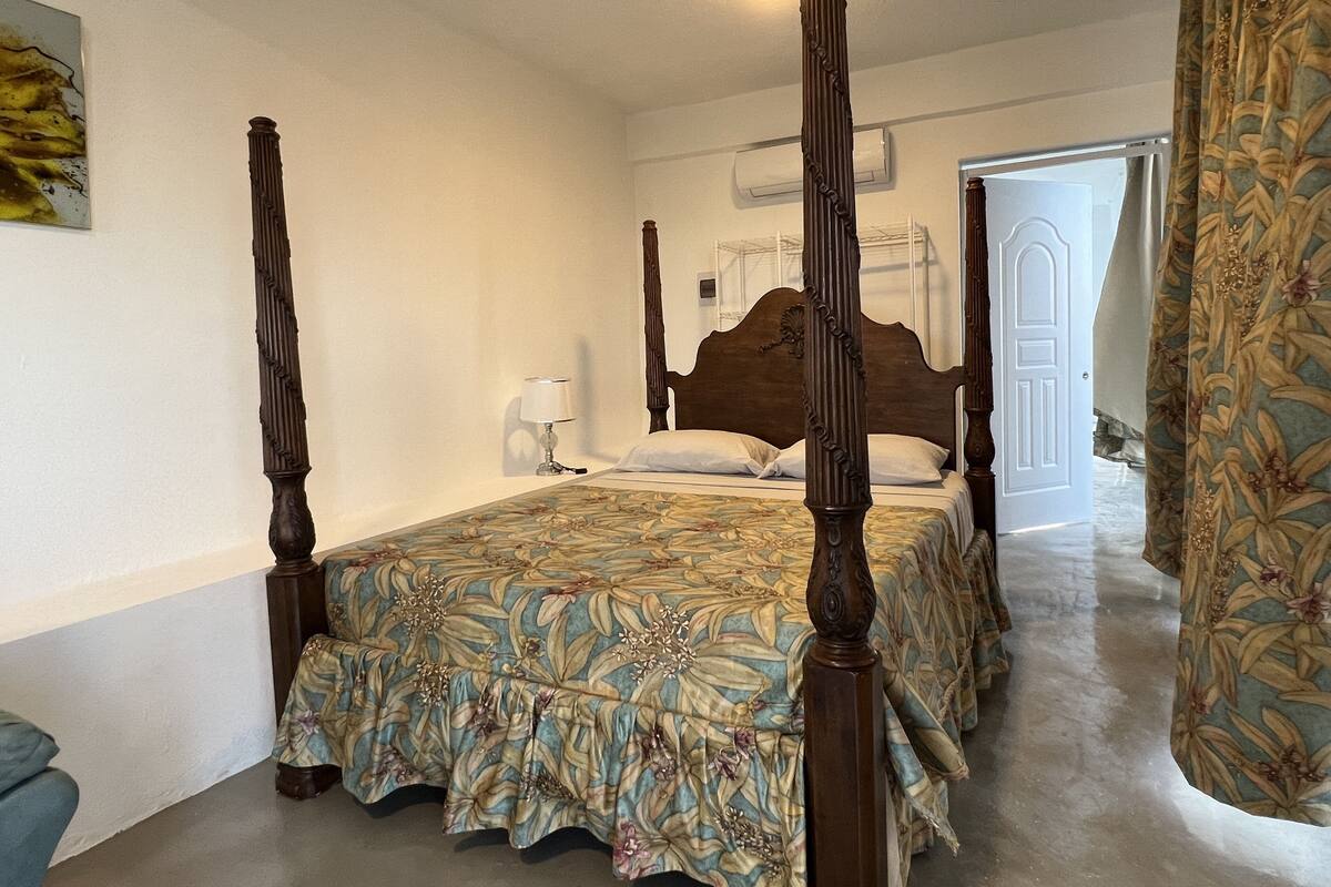 Una cama con dosel, ropa de cama floral, una mesita de noche con lámpara y una puerta que da a otra habitación.