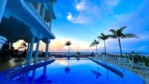 Zona de piscina con vista al océano, palmeras y un edificio con columnas.