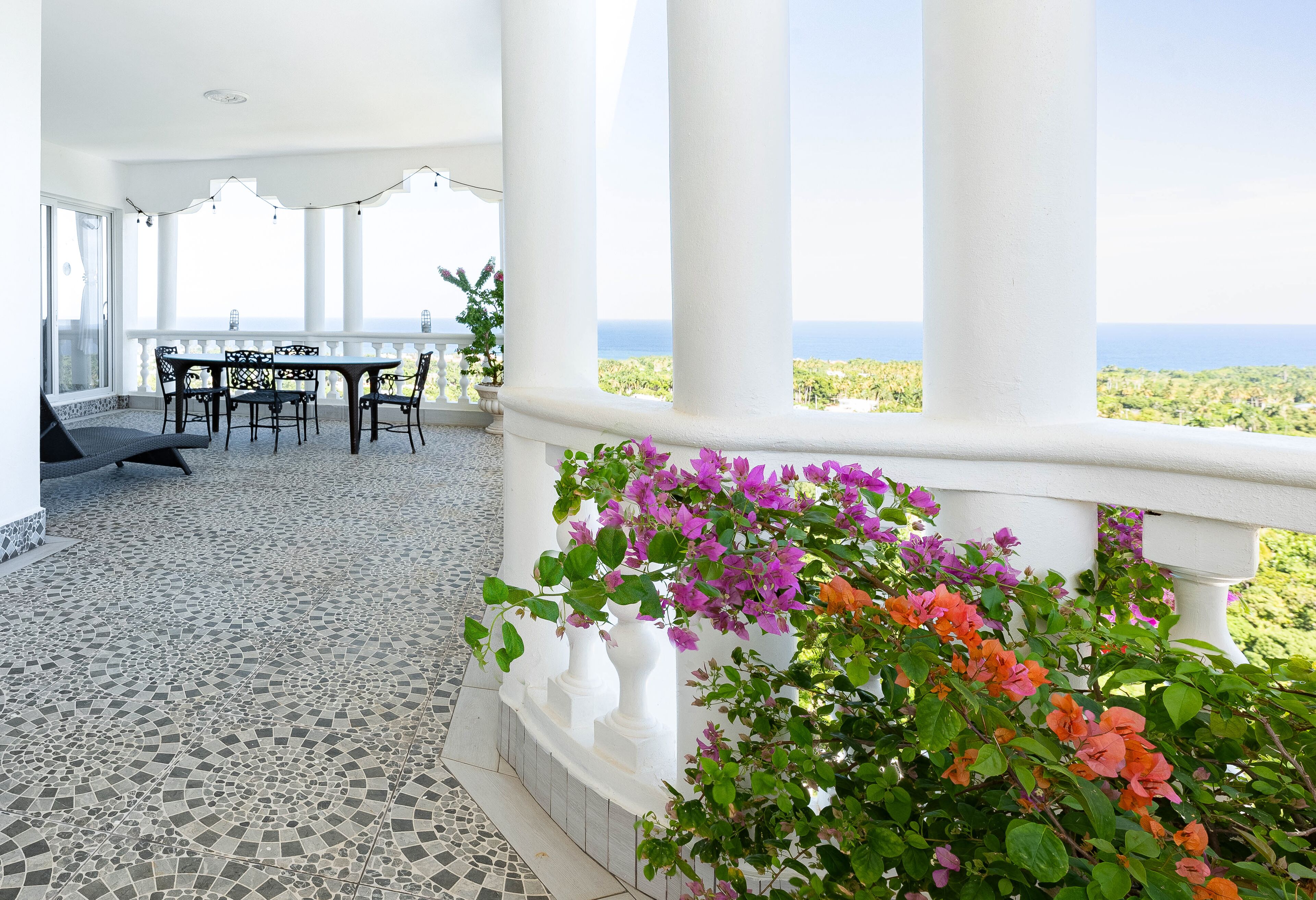 A covered outdoor area with a table and chairs, a white railing, and a view of the ocean.
