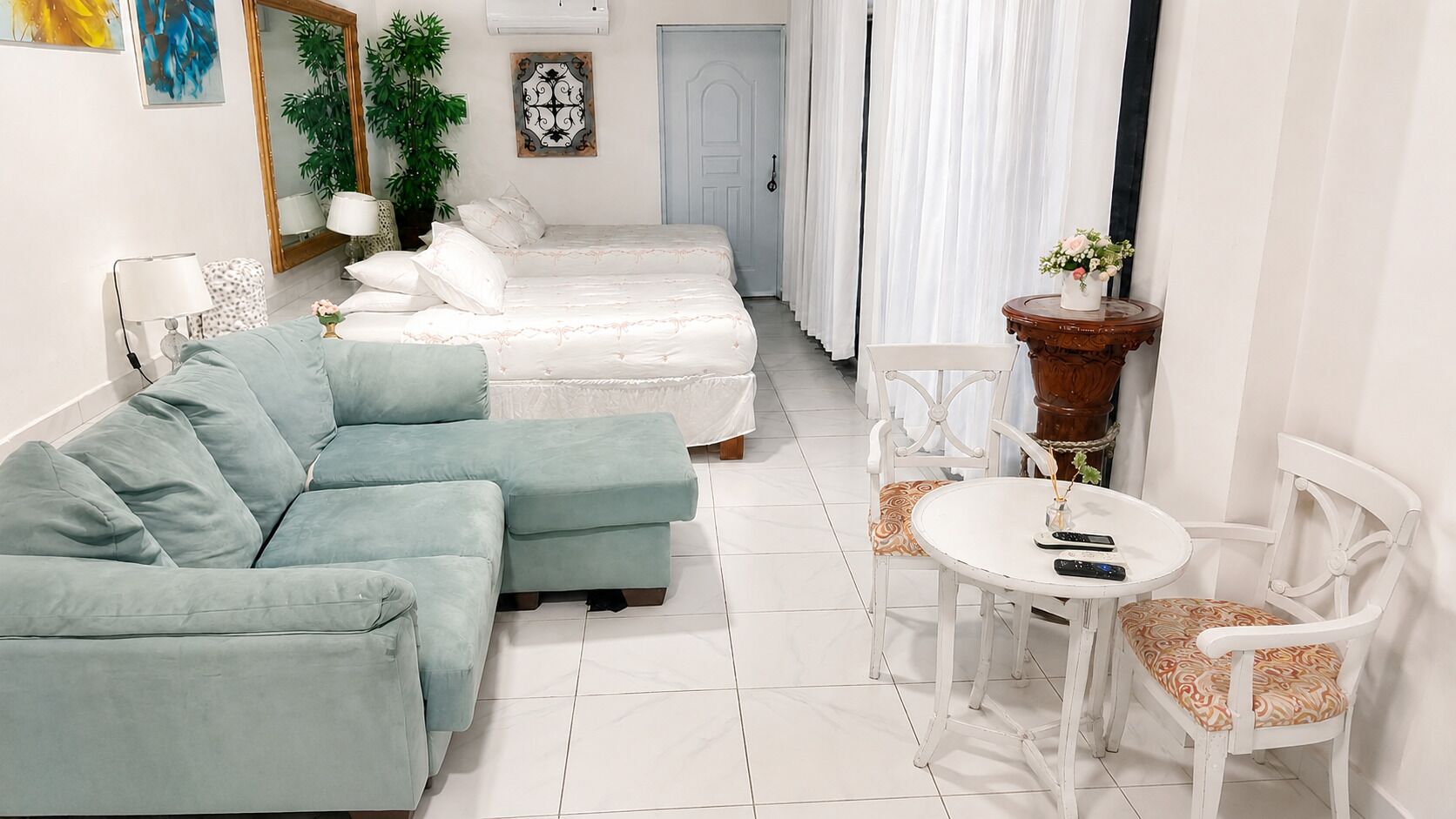 Una habitación con un sofá azul claro, una cama con ropa de cama blanca, una mesita redonda con sillas y una planta en maceta.