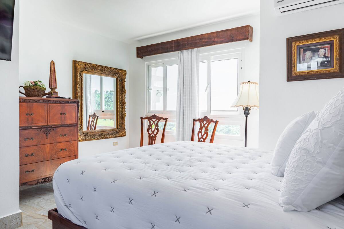 Un dormitorio con una cama grande, cómoda de madera, espejo decorado y una ventana con cortinas.