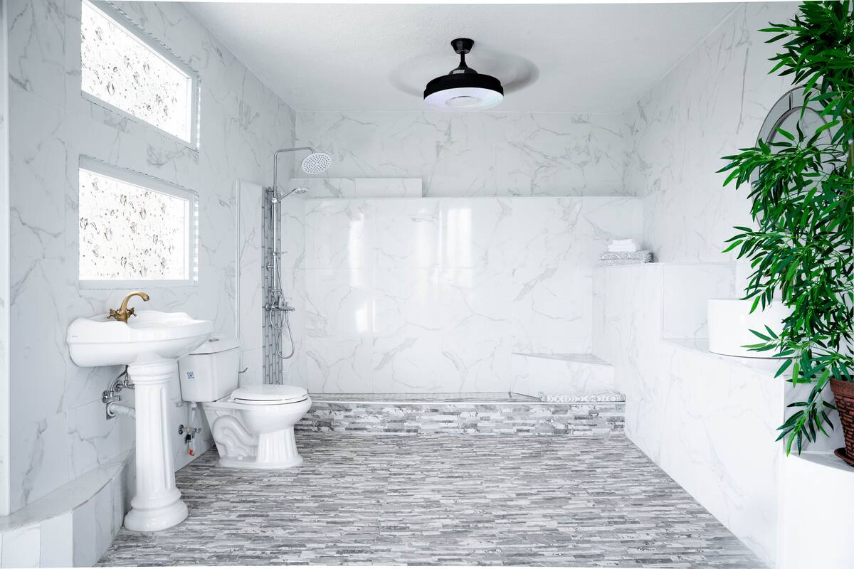 Un baño con suelo de mármol, lavamanos pedestal, inodoro, ducha y ventilador de techo.