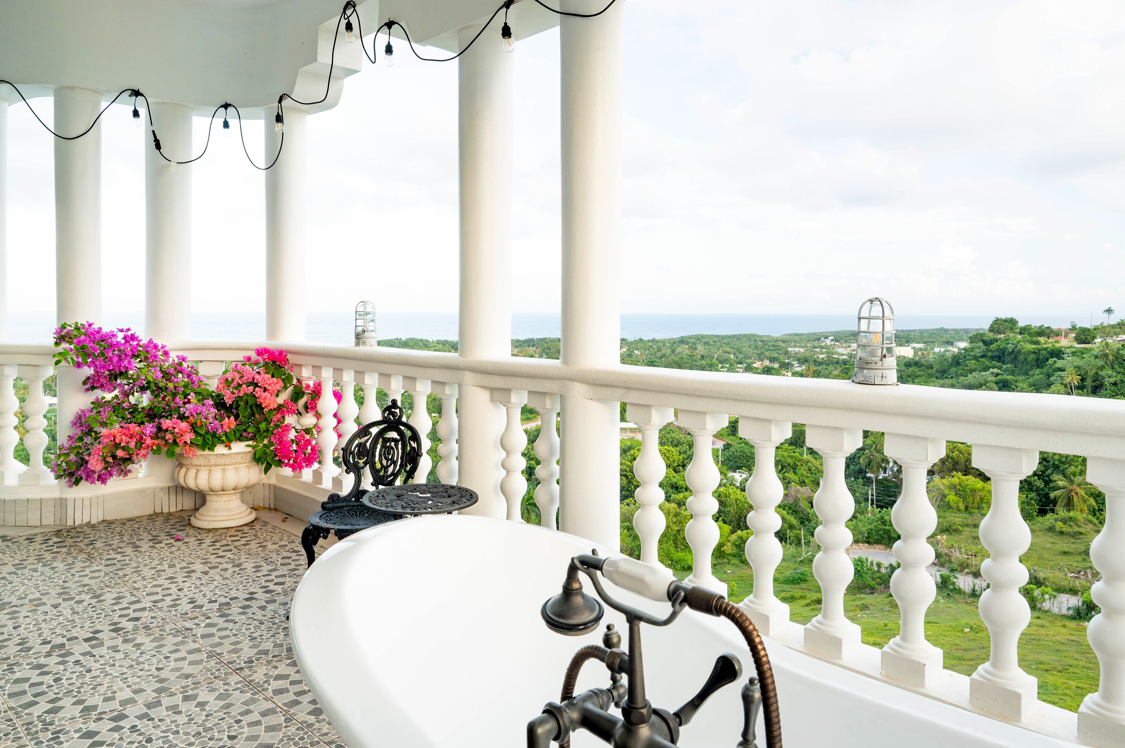 A balcony with a white bathtub, a potted plant with pink flowers, and a view of a green landscape.