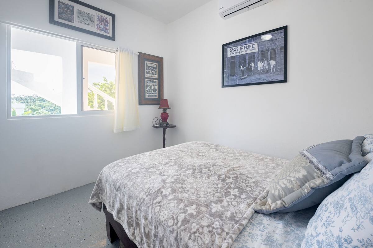 Un dormitorio con una cama, una ventana con cortinas, un cuadro enmarcado en la pared y una lámpara en una mesita de noche.