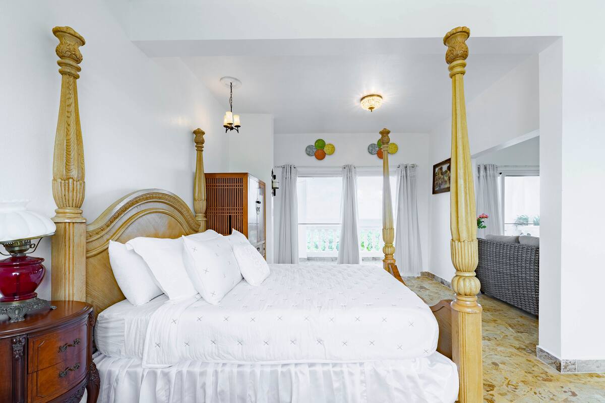 Una cama con dosel con sábanas blancas y cabecera de madera, una mesita de noche con una lámpara roja, y vistas a un balcón con cortinas.