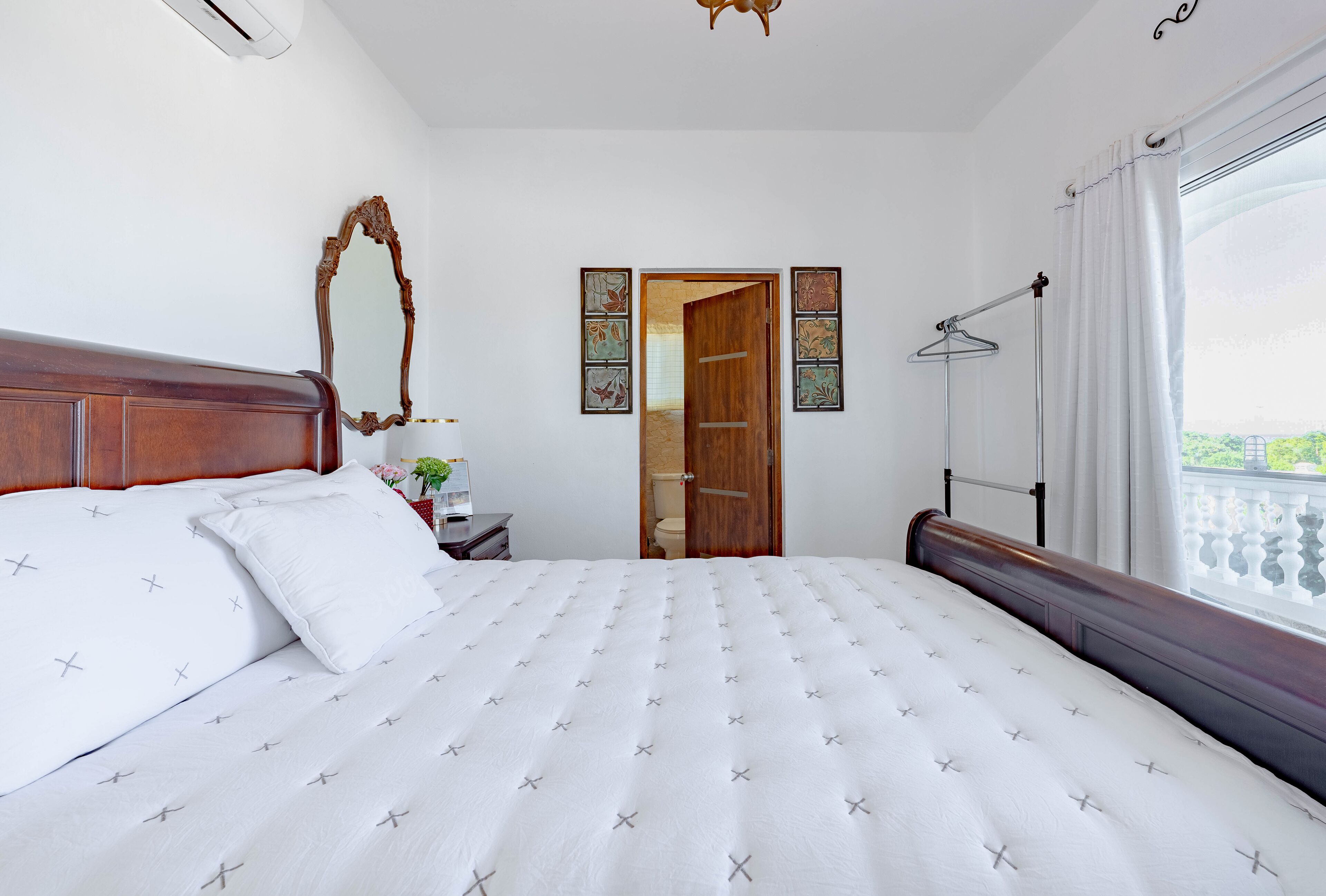A neatly made bed with a white bedspread and pillows, a wooden headboard, a mirror, and a window with a balcony view.