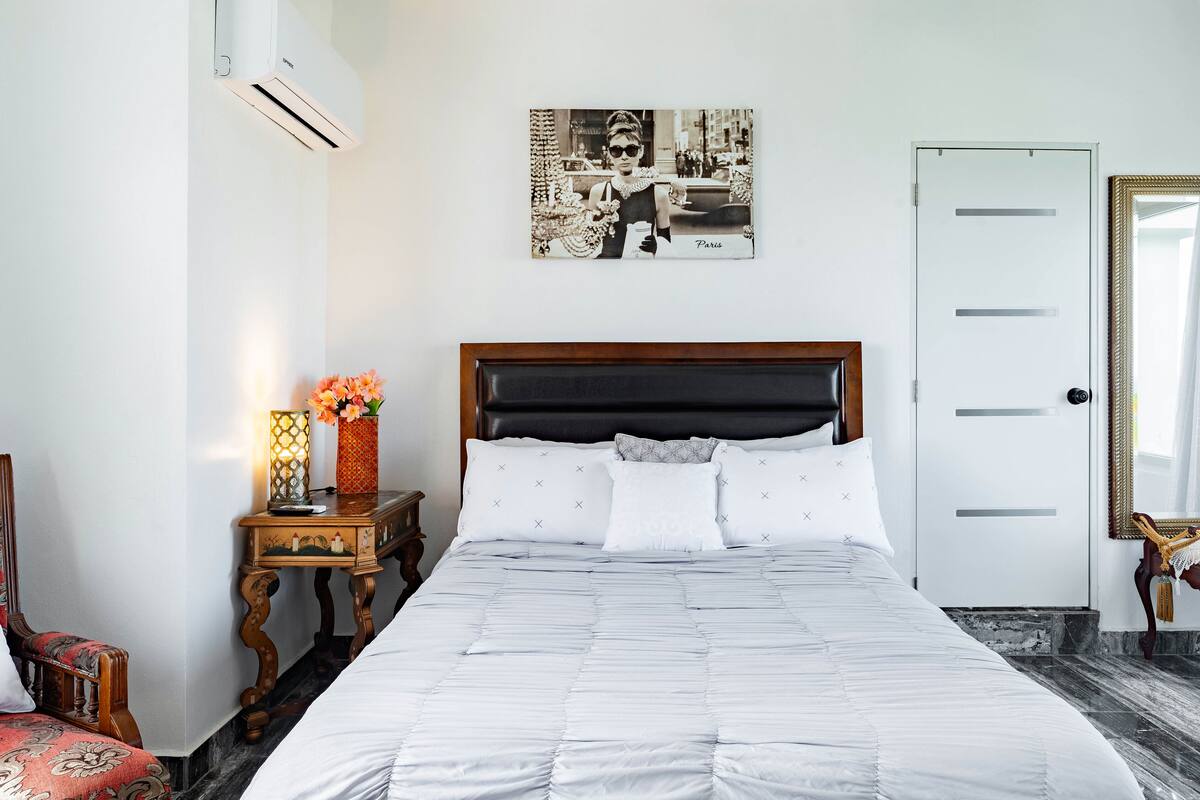 Una cama bien hecha con ropa de cama blanca, un cabecero de madera, una mesita de noche con una lámpara y un florero, y un cuadro en la pared.