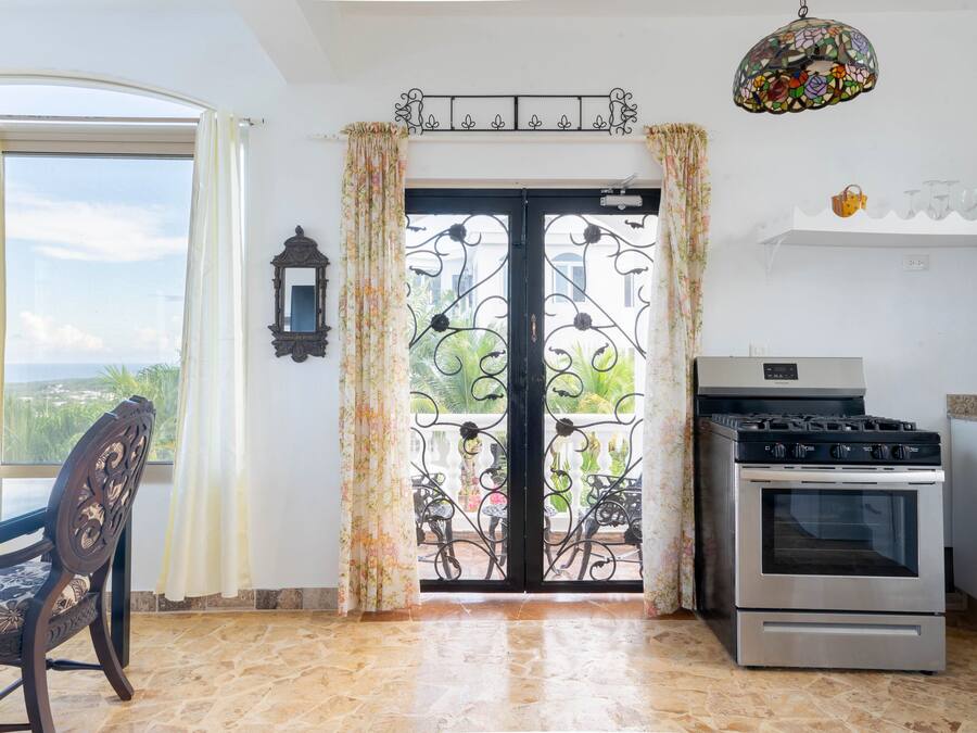 Una cuina amb forn, taula de menjador amb caires, finestra amb cortines i vista a verdura a l'exterior.