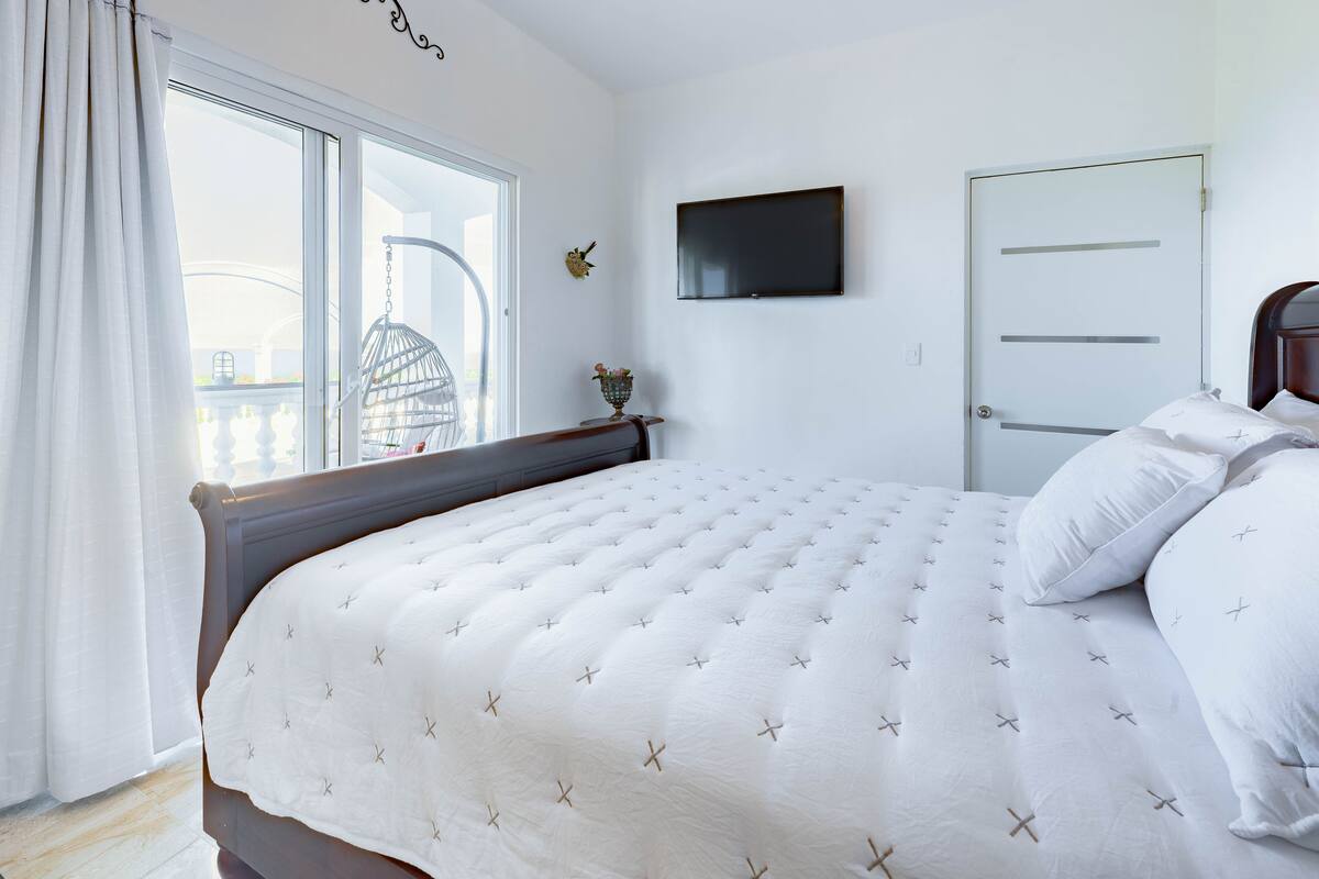 Un dormitorio con una cama grande, un televisor montado en la pared y una ventana con cortinas.
