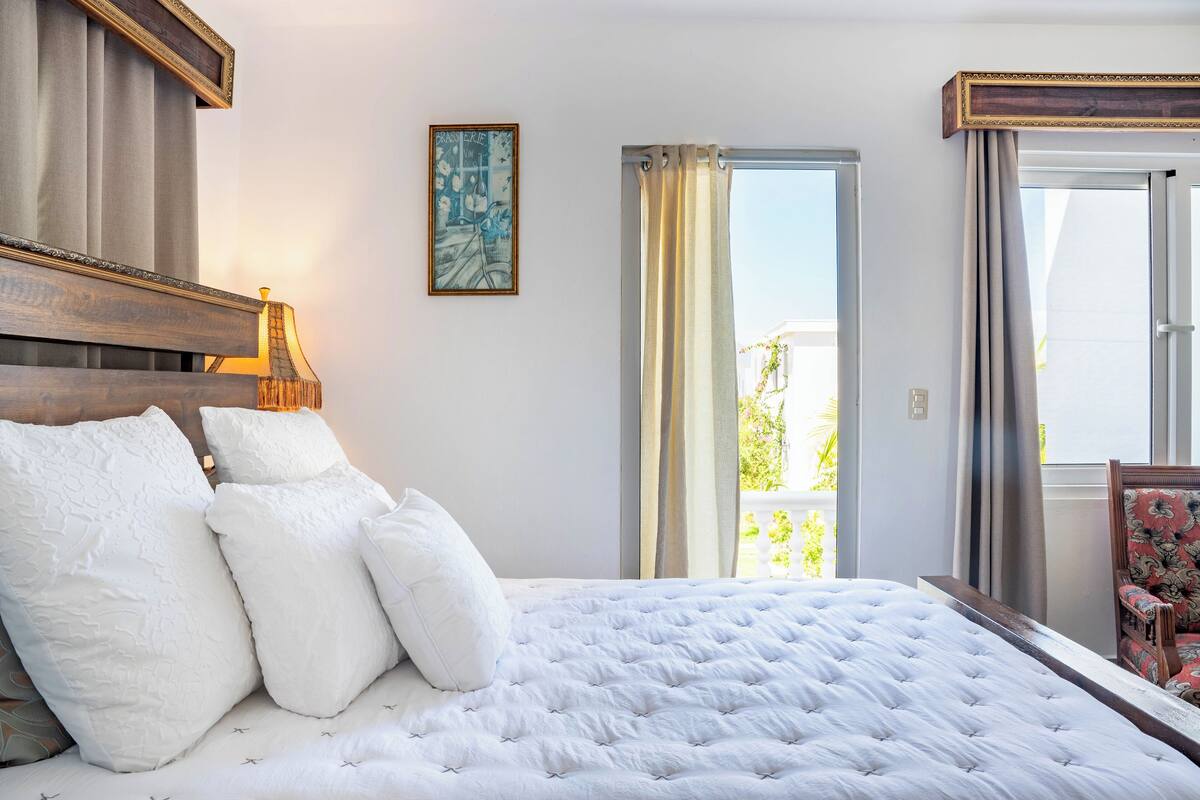 Un dormitorio con una cama grande, cabecera de madera, un cuadro enmarcado en la pared y una ventana con cortinas.