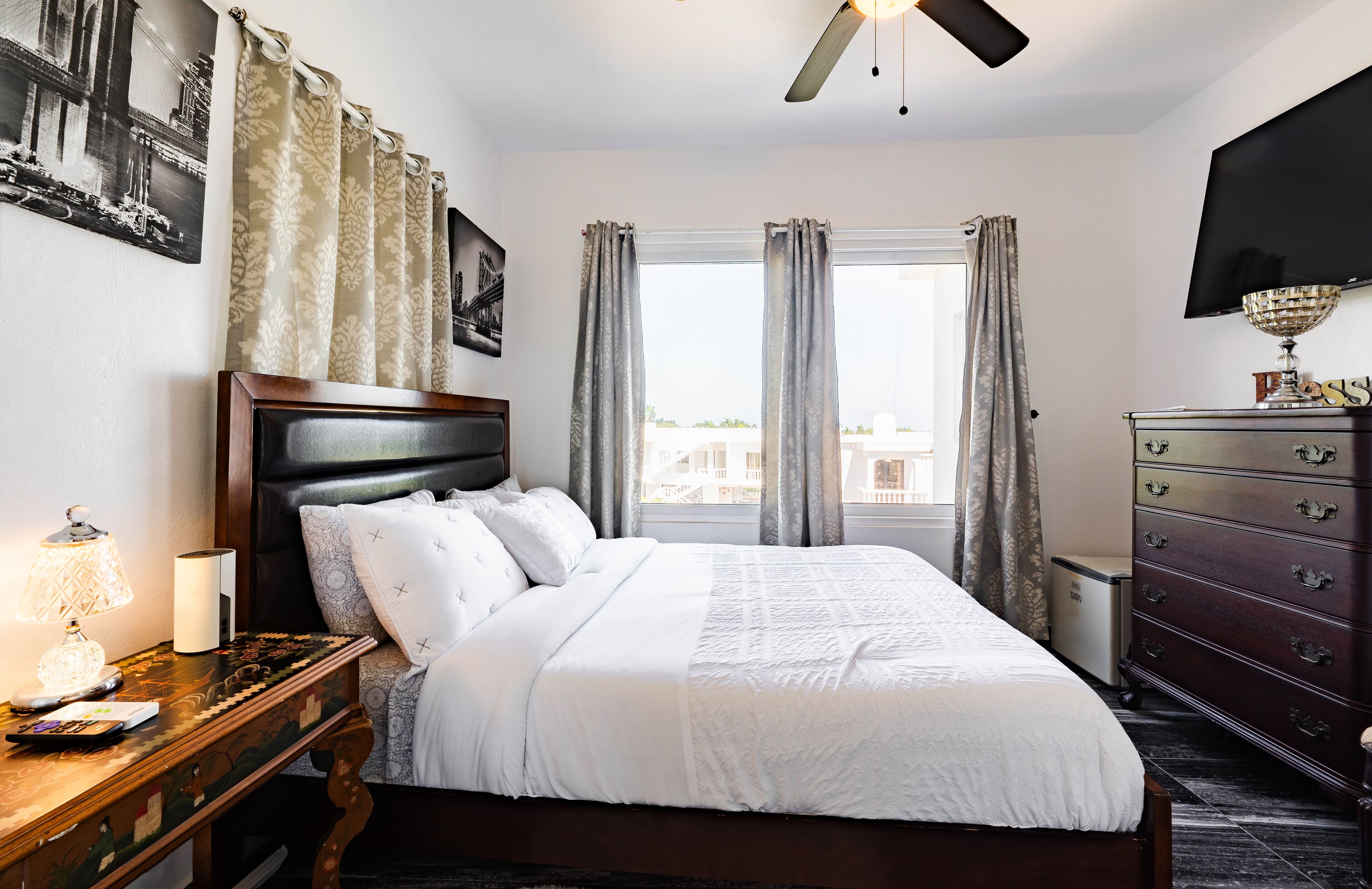 A neatly arranged bedroom with a bed, bedside table, lamp, dresser, and a television on the wall.