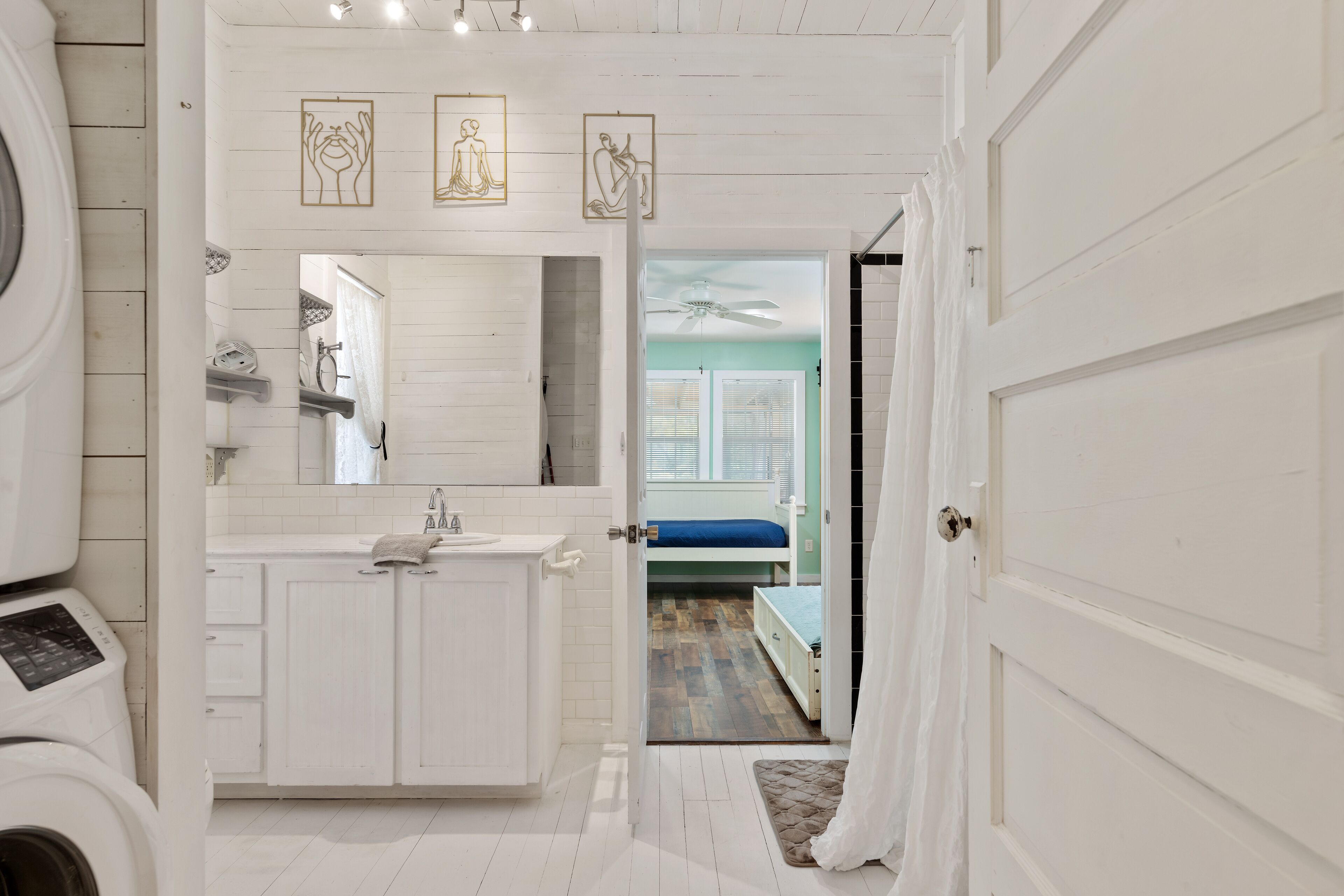 A bathroom with a white sink, mirror, and cabinets, leading to a bedroom with a bed and a ceiling fan.