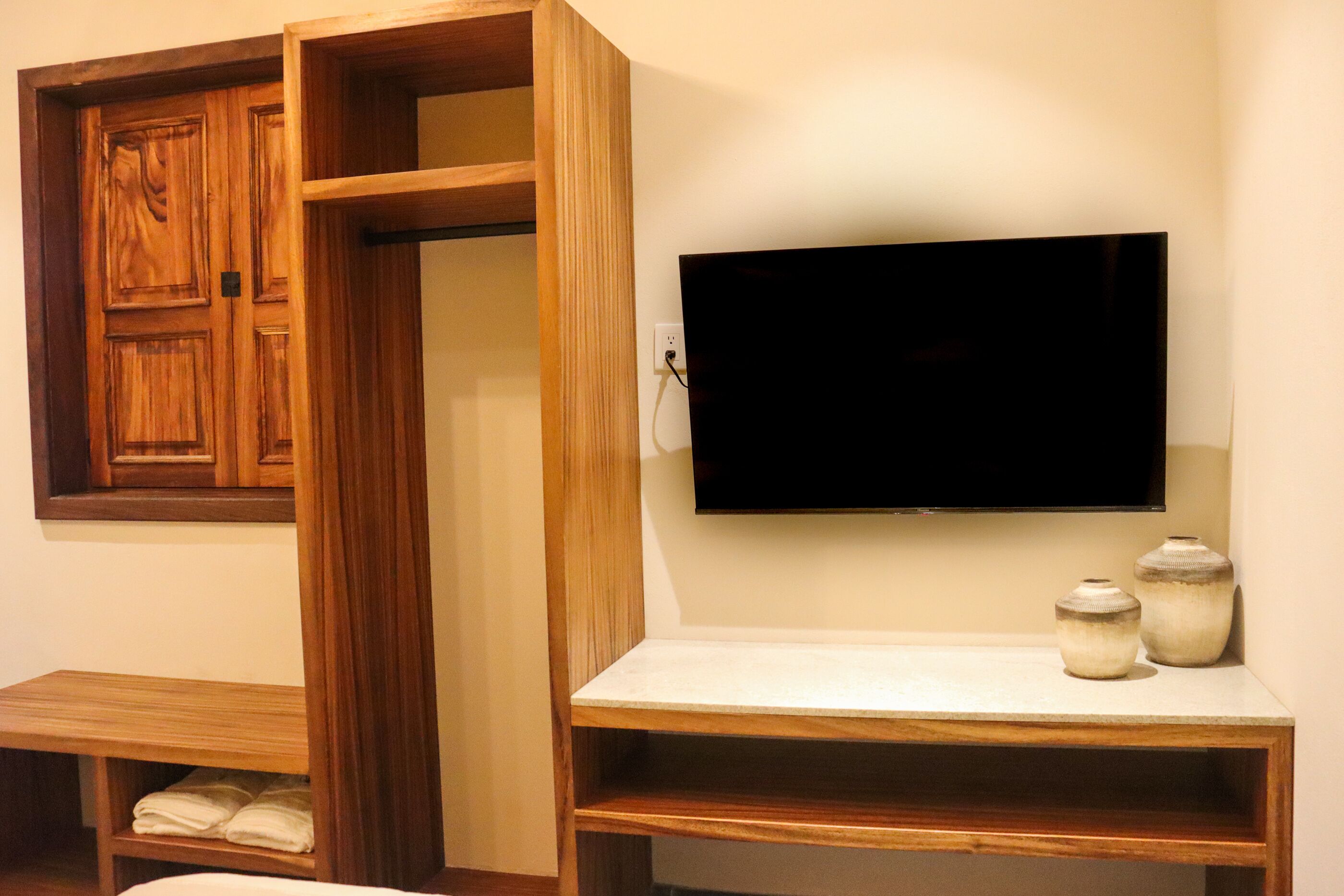 A room with a wooden wardrobe, a flat-screen TV mounted on the wall, a small wooden table, and two ceramic jars on the table.