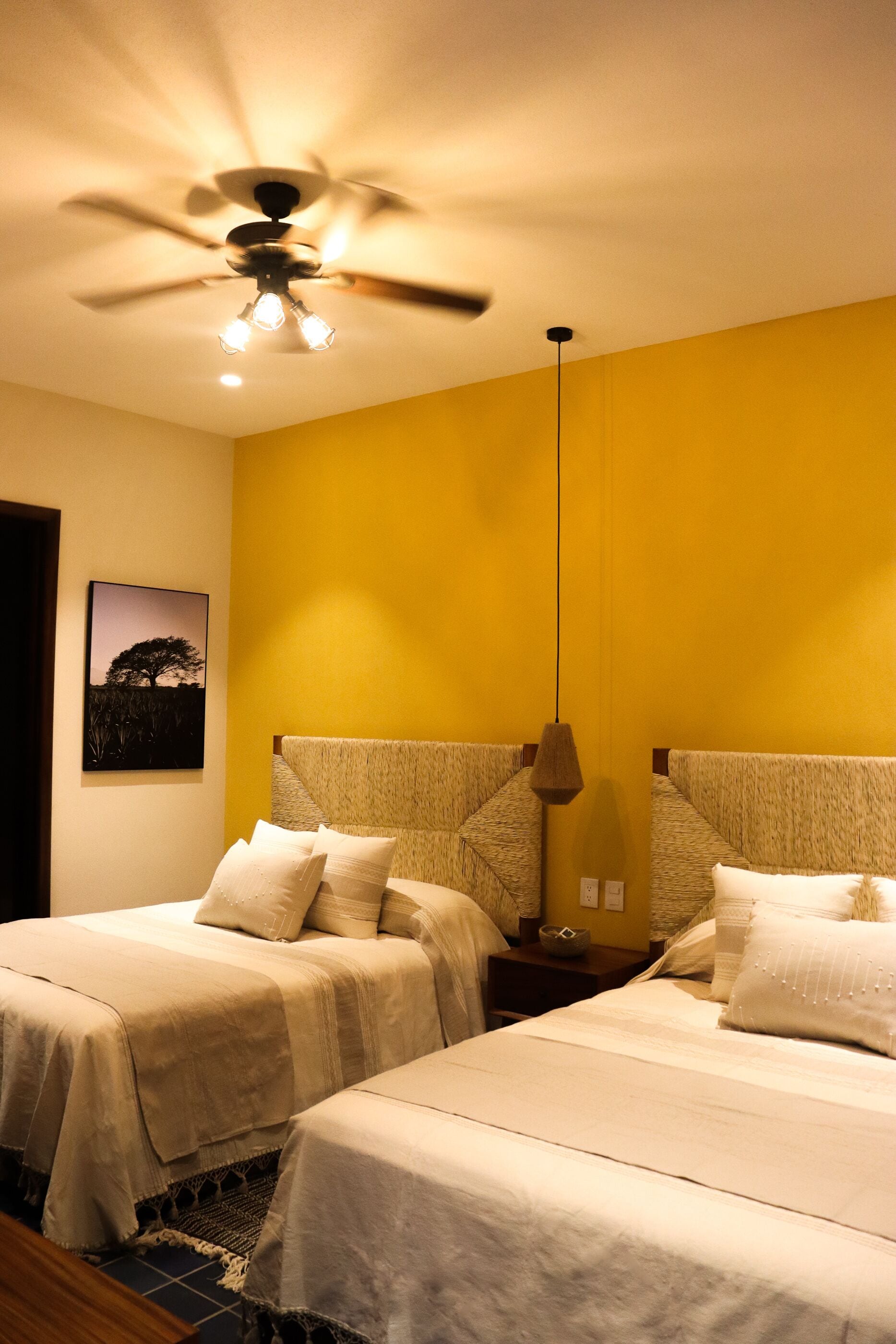 Quarto de hotel com duas camas, um ventilador de teto e luminária fixada na parede.