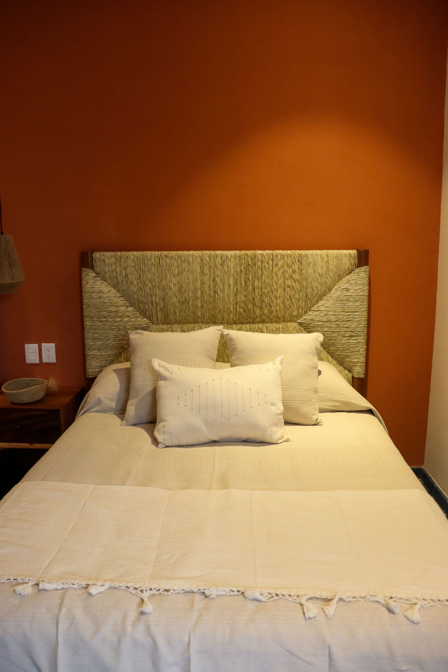 A neatly made bed with a textured headboard, a bedside table with a basket, and a wall socket.