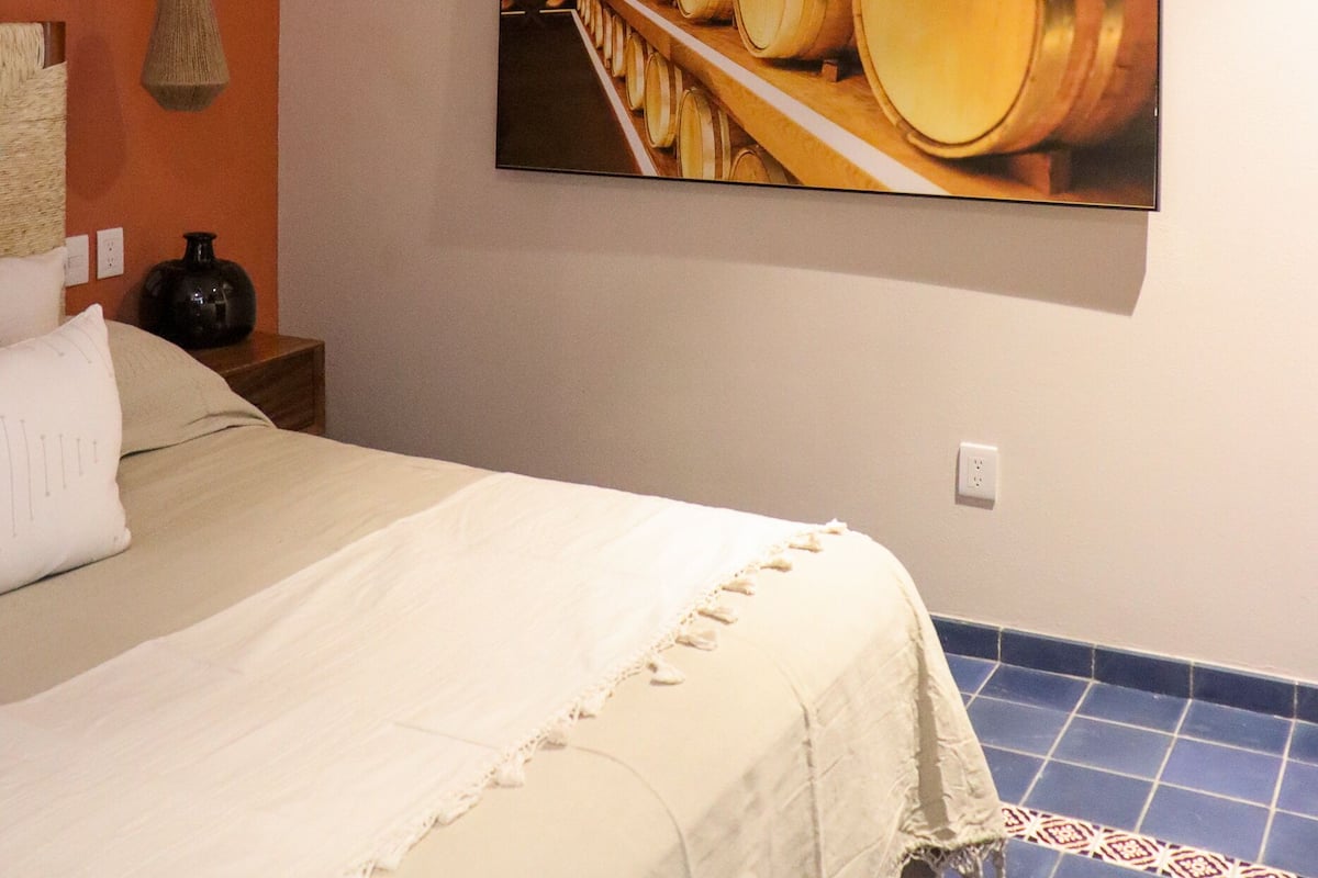 A hotel room with a bed, a ceiling fan, an air conditioner, and a wall-mounted painting of barrels.