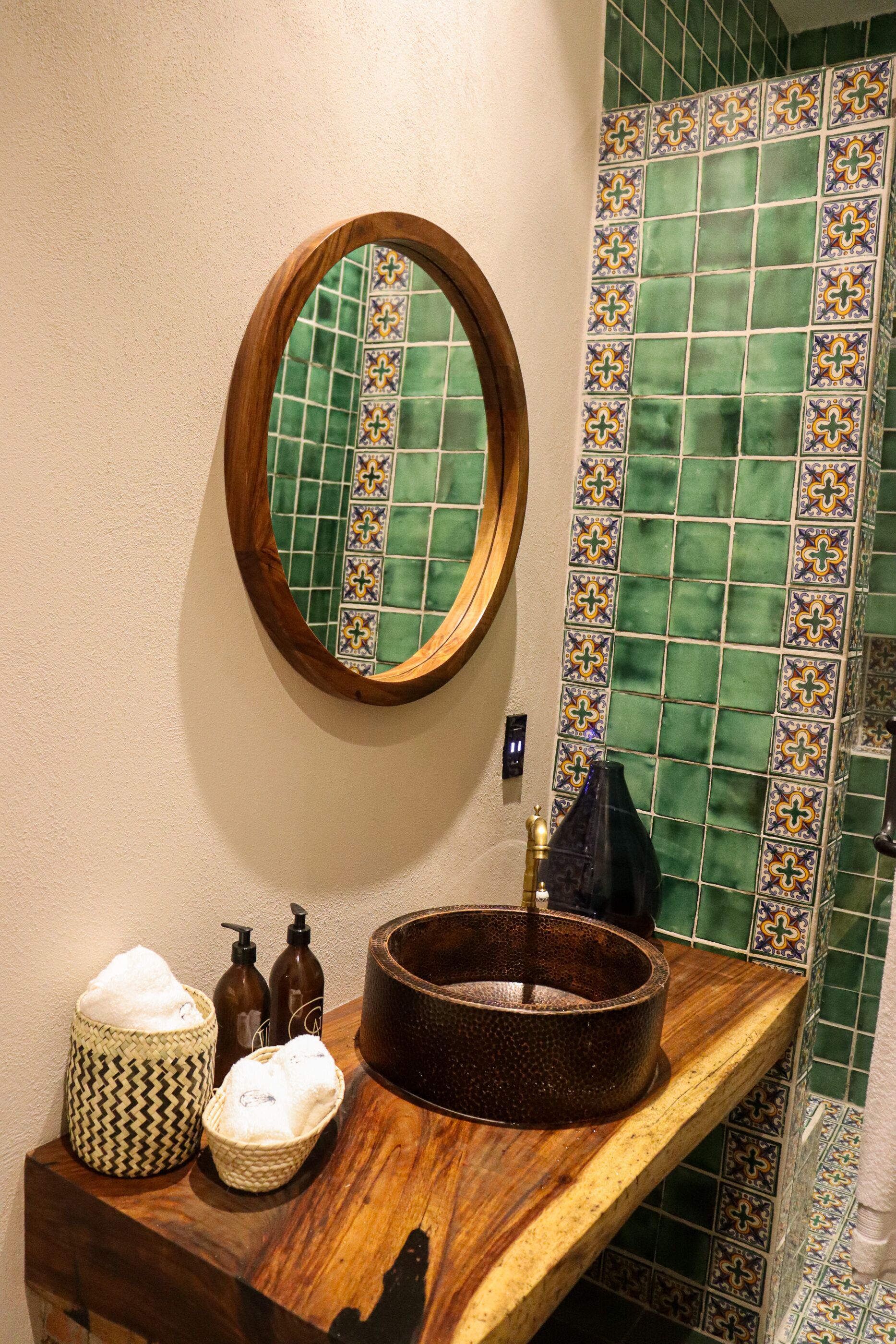 A bathroom with a wooden vanity, a round mirror, and a green tiled backsplash.
