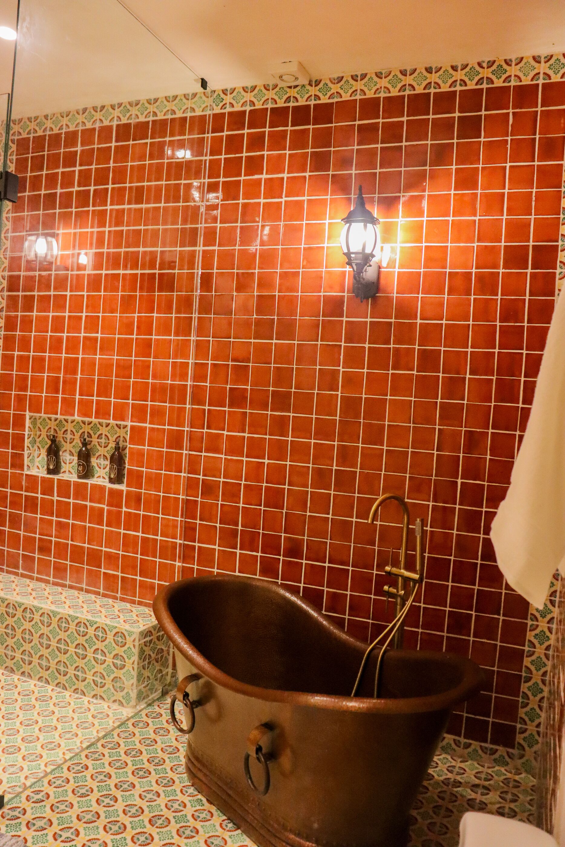 A bathroom with a large copper bathtub, patterned tiles, and a wall-mounted light fixture.