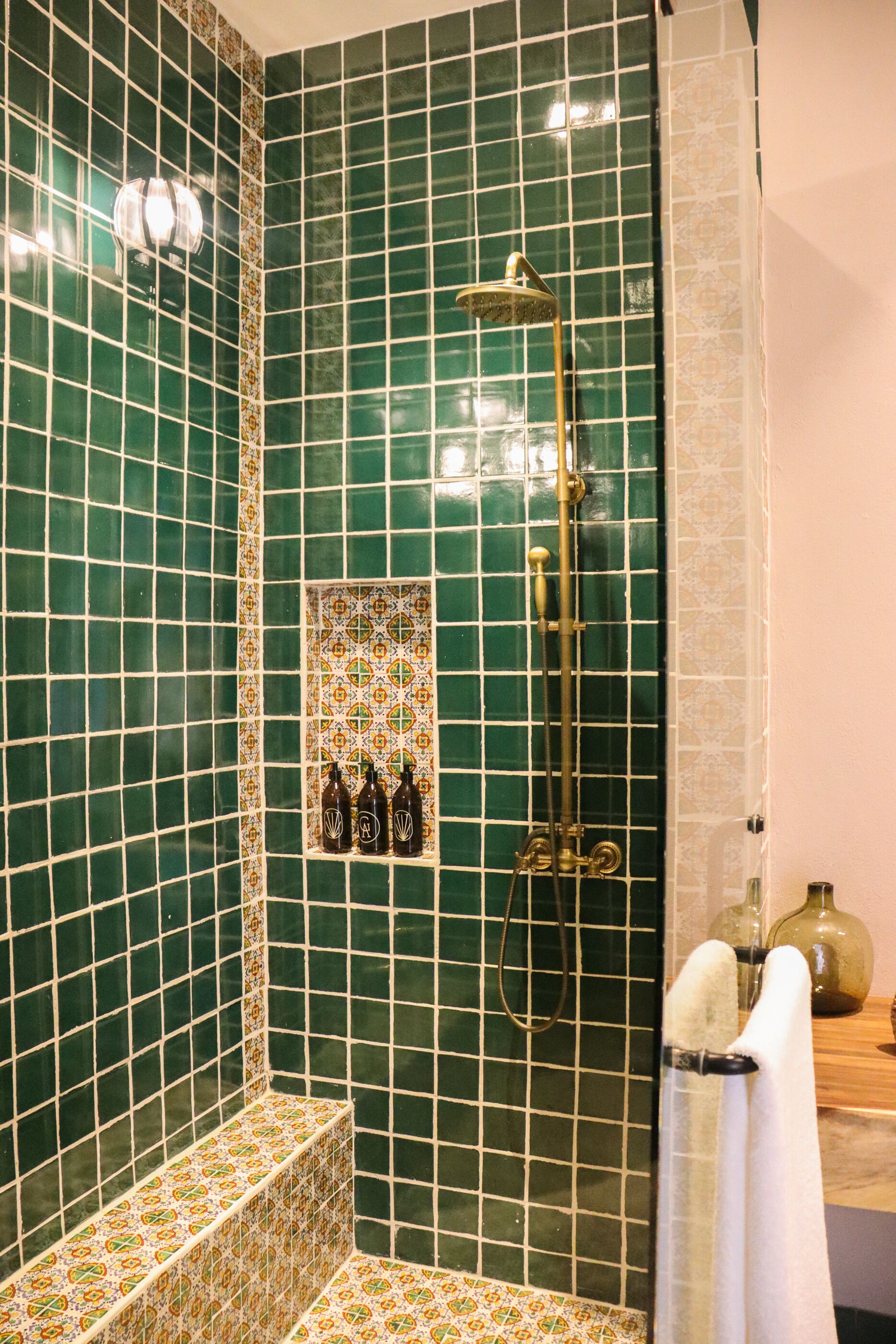 A bathroom with green tiled walls, a shower with a golden showerhead, and a small shelf with bottles.