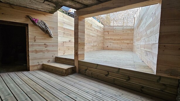 Una baita di legno con un'ampia finestra, un terrazza in legno e una piccola scala che conduce a una piattaforma.