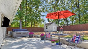 A patio with a metal table, chairs, and a red umbrella. There are potted plants and a small shed in the background. - The Sophey (Denison)