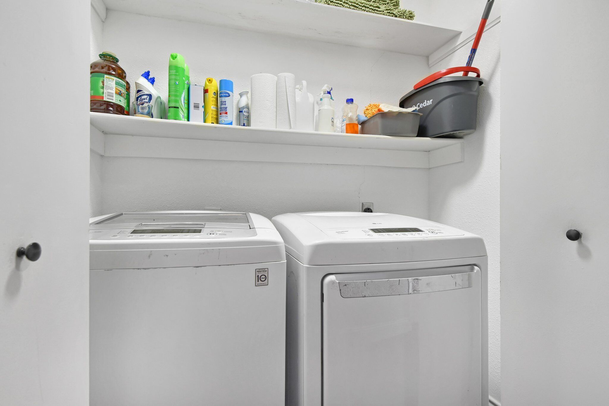 Une buanderie avec deux machines à laver blanches et une étagère au-dessus, où sont rangés les produits de nettoyage.