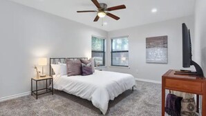 A bedroom with a bed, a ceiling fan, a nightstand, a TV, and a window with blinds. - The Sophey (Denison)