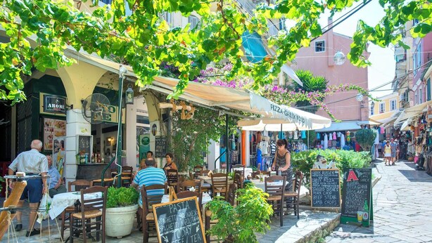 Café al aire libre con mesas y sillas, rodeado de edificios y vegetación.