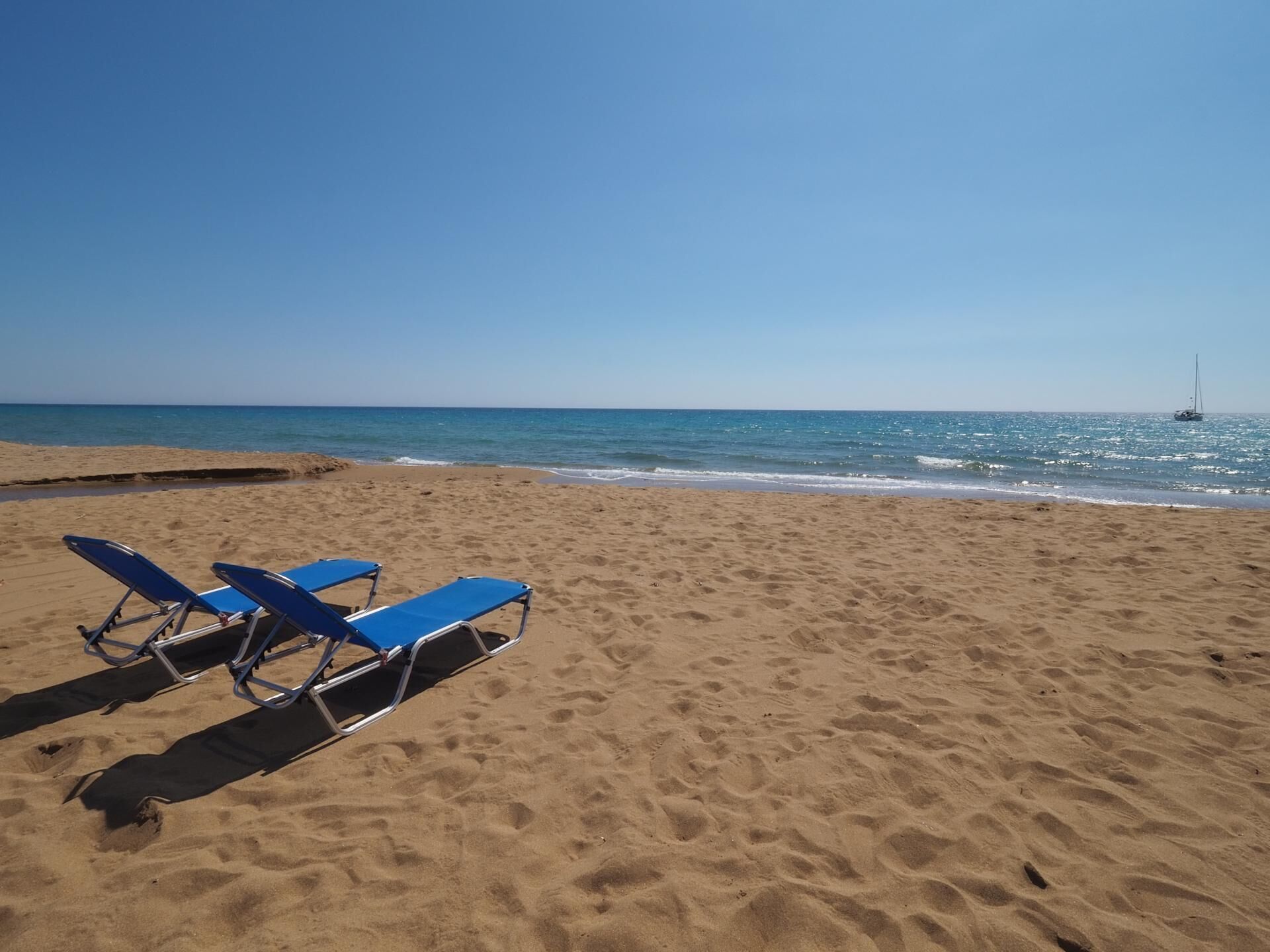 A beach with two blue lounge chairs and a sailboat in the distance.