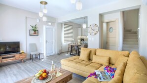 Una sala de estar moderna con sofá, mesa de centro y televisor. Se observa un comedor con mesa y sillas, y una cocina al fondo.