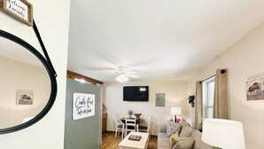 A cozy living room with a sofa, coffee table, and a TV mounted on the wall. There is a dining area with a table and chairs, and a kitchen visible in the background.