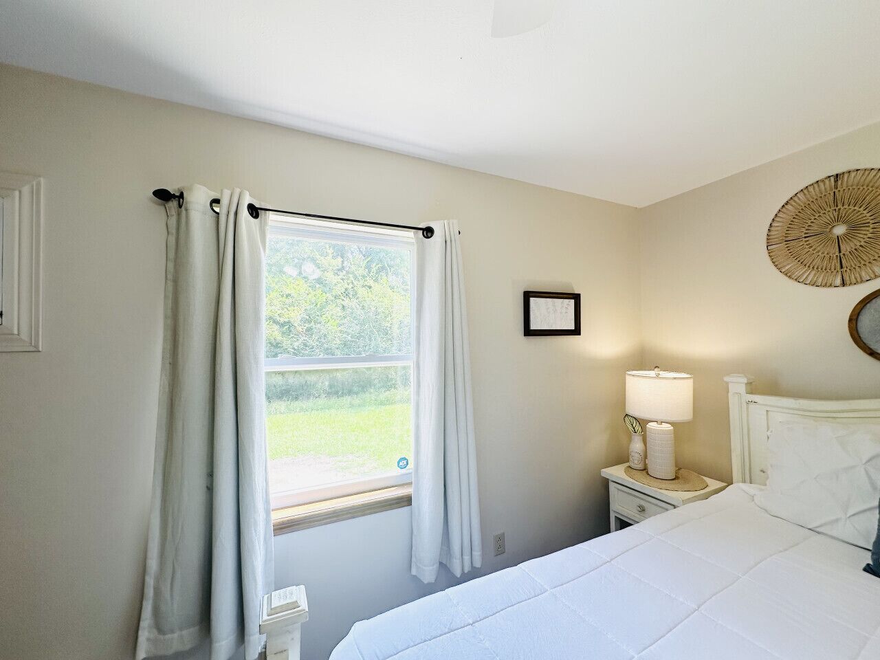 Une chambre à coucher comprenant un lit, une table de chevet avec une lampe, une fenêtre avec des rideaux et un élément décoratif fixé au mur.