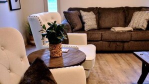 Una sala de estar con un sillón blanco, un sofá marrón, una mesa de centro de madera y una mesita auxiliar con una planta.
