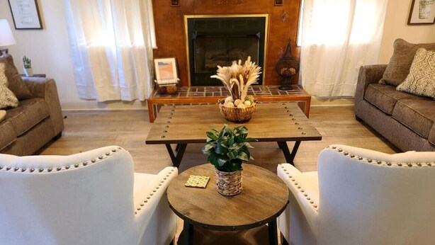 Una sala de estar con chimenea, dos sillones blancos, una mesa de centro de madera con una planta y una cesta con objetos decorativos.
