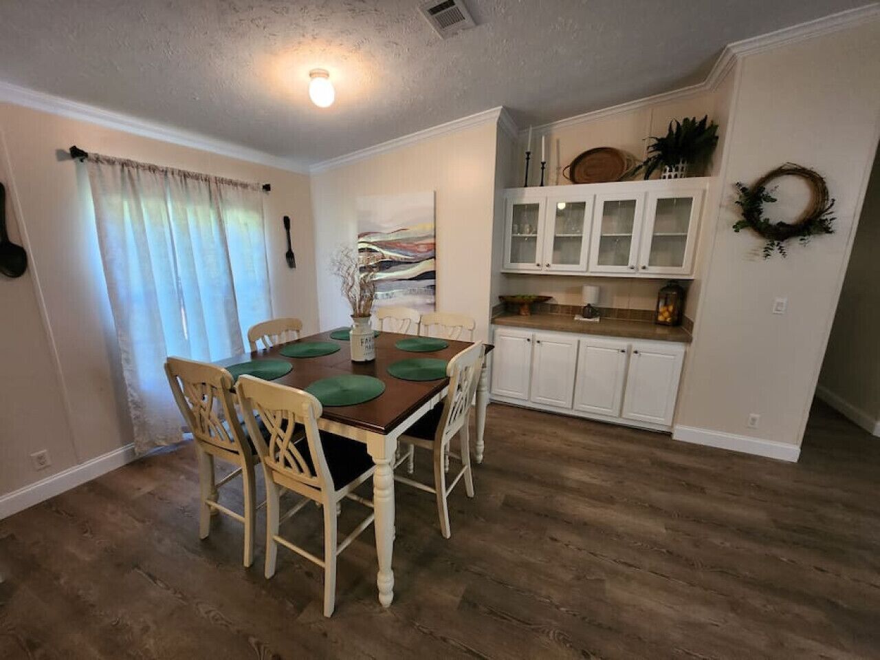 Uma sala de jantar com mesa e cadeiras de madeira, uma cozinha com armários brancos e uma janela com cortinas.