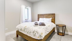 A bedroom with a wooden bed, white bedding, a round side table, and a window with white curtains. - Unique Grand Junction Guest Suite w/ Gas Fire Pit! (Grand Junction)