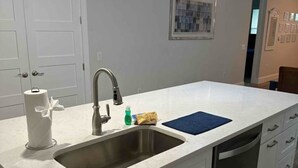 A kitchen with a white countertop, stainless steel sink, and a dishwasher. There are three pendant lights hanging above the sink. - Delightful 3-bedroom house in Bradenton with WiFi, Games, and more #2 of 2 (Bradenton)