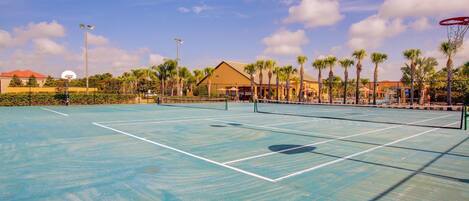 Una cancha de tenis al aire libre con red, rodeada de palmeras y un edificio de techo rojo.