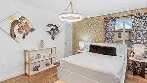 Une chambre à coucher avec un lit, une console blanche agrémentée d’objets décoratifs, une œuvre d’art accrochée au mur, un rideau à motifs et une fenêtre avec des stores.