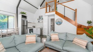 Uma sala de estar moderna com um sofá cinzento, uma escada de madeira e uma cozinha ao fundo.