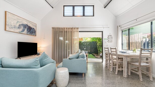 Uma sala de estar moderna com televisão de tela plana, dois sofás azul-claros, uma área de jantar com mesa e cadeiras de madeira, e vista para um jardim através de uma porta deslizante.